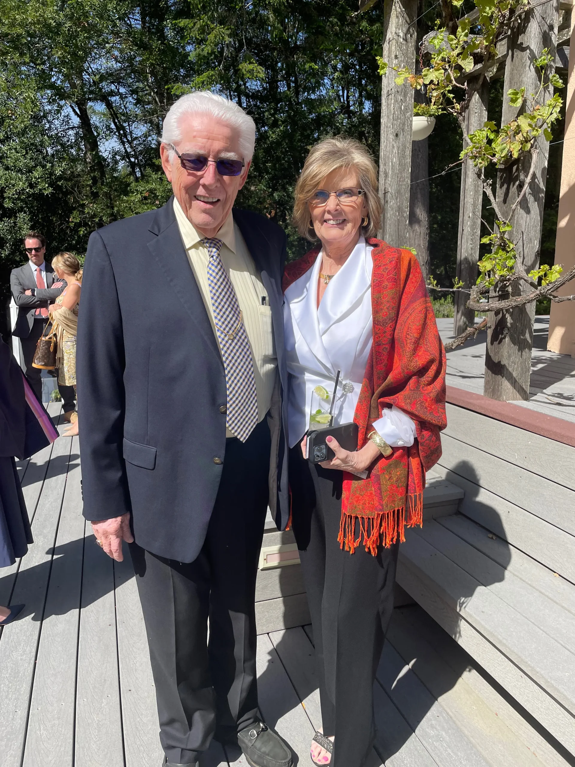 An elderly couple posing together on a wooden deck outdoors, dressed in formal attire, smiling at the camera.